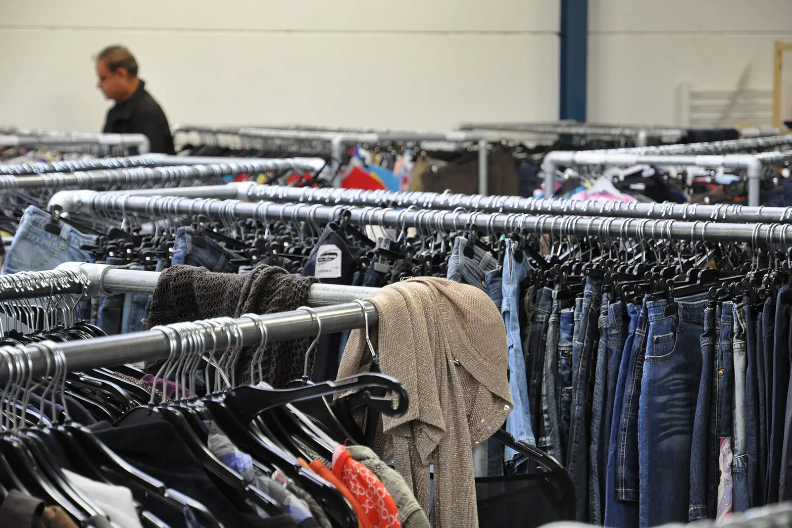 Packed rails of secondhand clothing in a charity shop, with a shopper browsing behind them