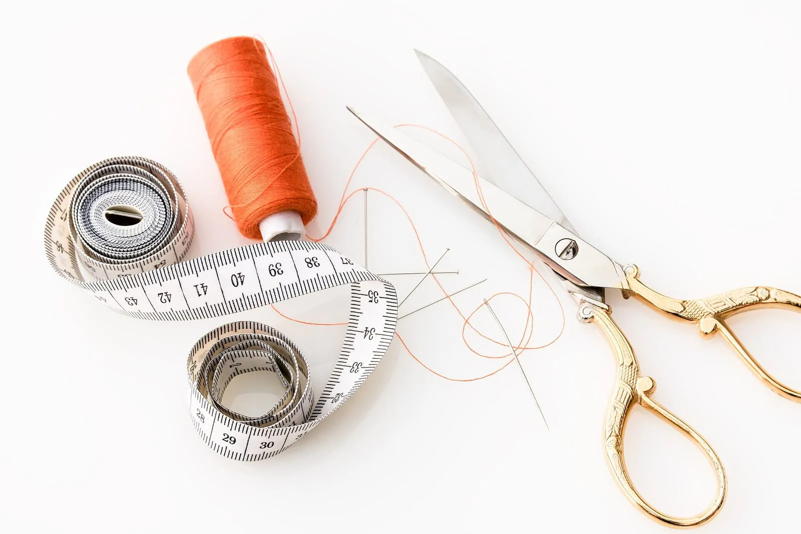 A coiled soft tape measure, orange thread, pins and dressmaking scissors on a white surface